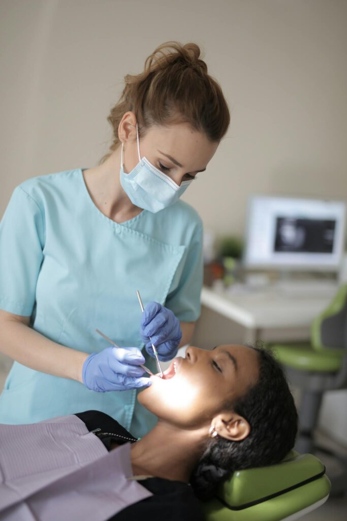 Side view of young female dentist in medical mask and latex gloves wearing blue uniform holding medical instruments while treating teeth of female ethnic patient in modern clinic